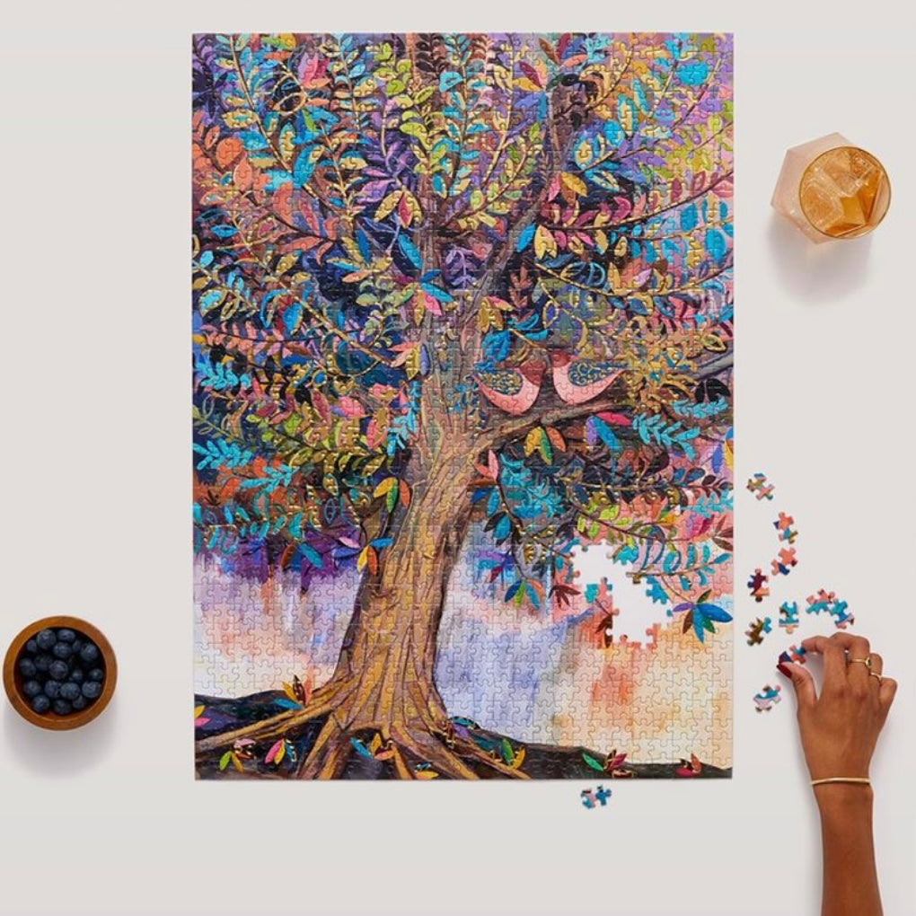 Assembled colorful jigsaw puzzle depicting a tree with gold foil embellishments, surrounded by a puzzle piece border, with a person's hand placing a piece on the tree.
