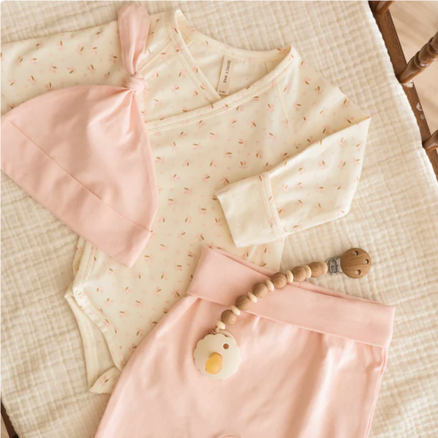 Baby outfit with pink romper, bib, and wooden teether on a textured surface