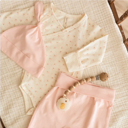 Baby outfit with pink romper, bib, and wooden teether on a textured surface