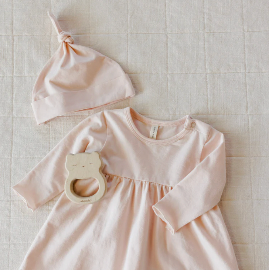 Pink baby dress with long sleeves, matching hat, and wooden teething ring on a textured beige surface.