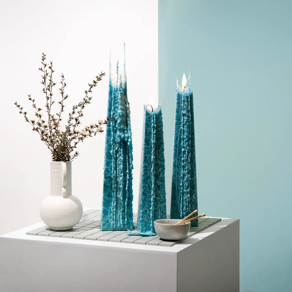Three tall blue candles on a white surface with a light blue wall in the background.
