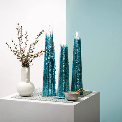 Three tall blue candles on a white surface with a light blue wall in the background.