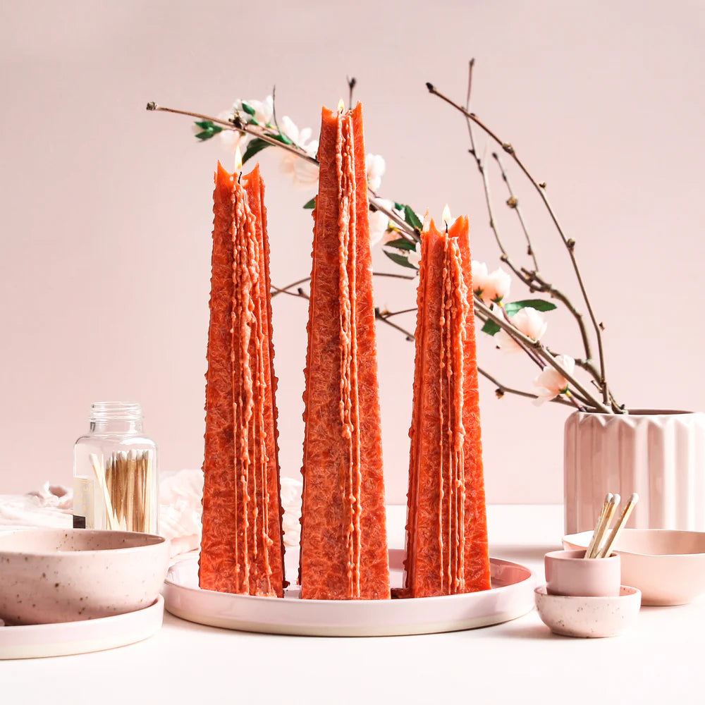Three tall, orange candles on a white plate with decorative branches and bowls in the background.
