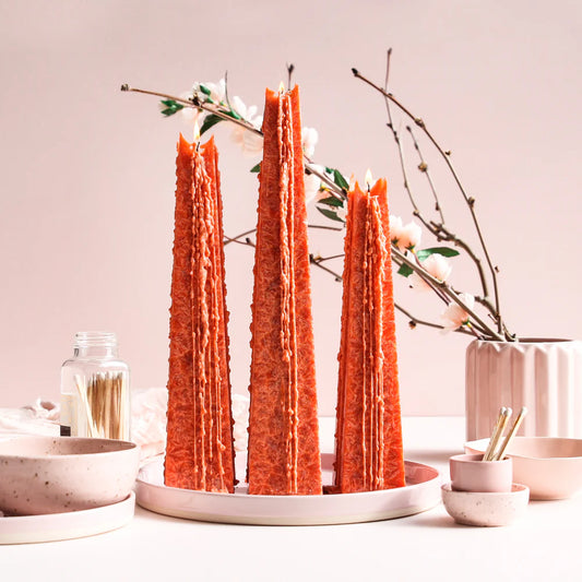 Three tall, orange candles on a white plate with decorative branches and bowls in the background.