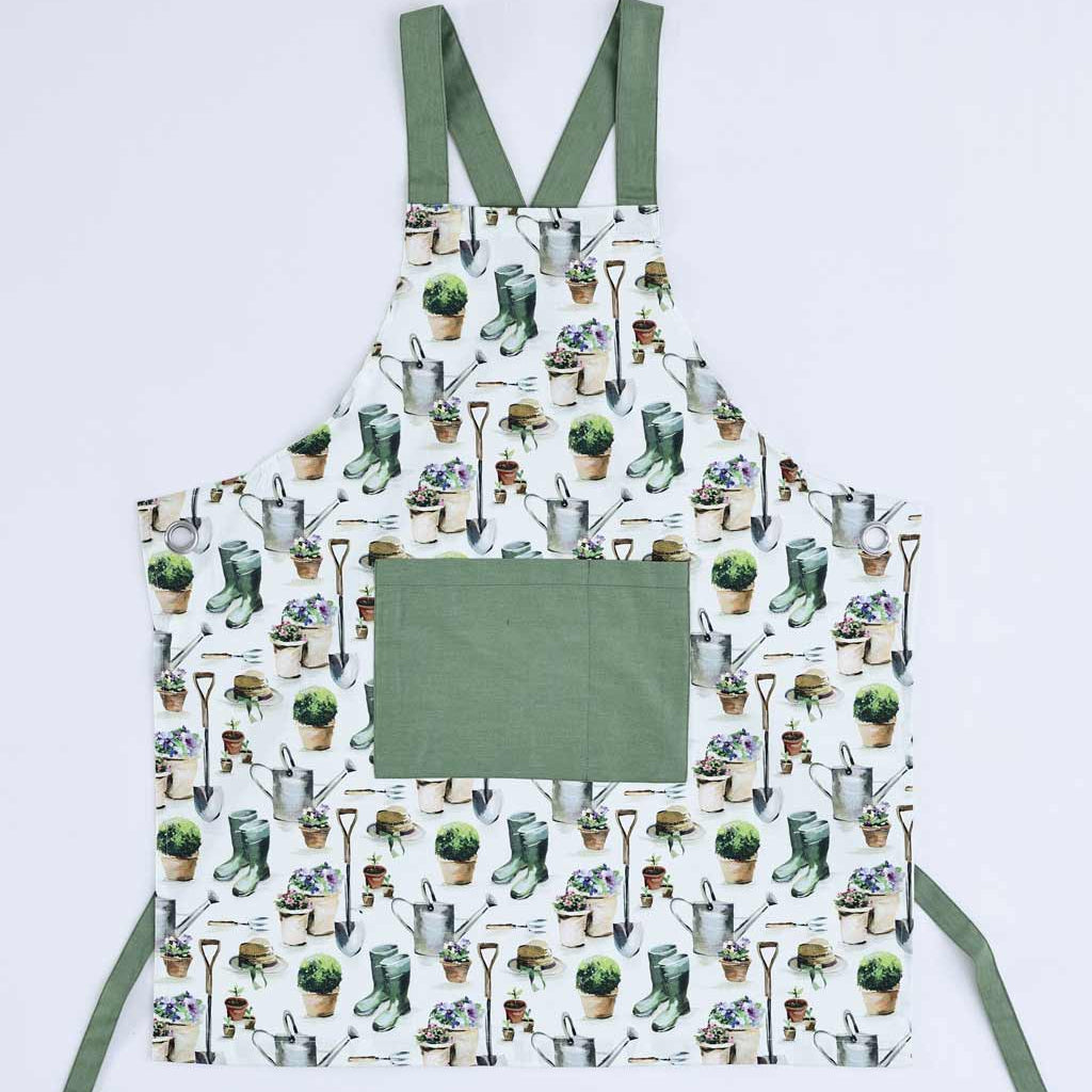A white apron with an all-over gardening-themed print of pots, plants, gloves, and tools, featuring solid sage green straps and a large solid sage green front pocket. The apron is laid flat against a light gray background.