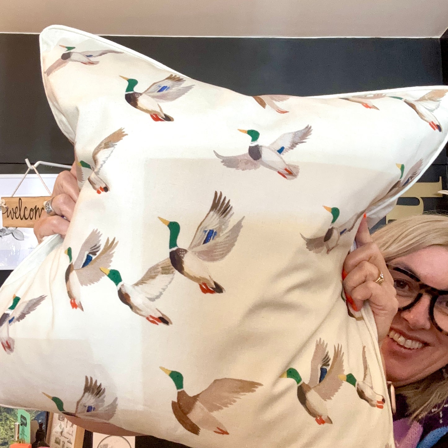 A person holding up a square cushion with a pattern of mallard ducks in flight on a beige background.
