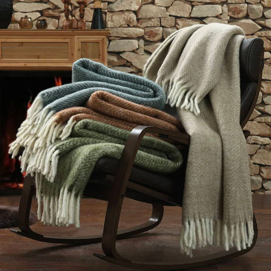 A collection of folded New Zealand wool throw blankets in various colors, including green, brown, beige, and blue, displayed on a wooden rocking chair in front of a stone fireplace.