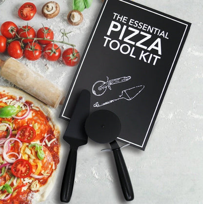 A pizza tool kit set arranged on a light stone surface with pizza ingredients around it. The kit includes a black rectangular box labeled 'THE ESSENTIAL PIZZA TOOL KIT,' a black pizza cutter, a black spatula, and a black server.