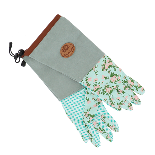 A pair of gardening gloves with a sage green canvas back, brown trim, a drawstring, and a round leather logo patch. The palms and fingers are covered in two different fabric patterns: one with a light green and white polka dot print, and the other with a pink and green floral print on a mint green background.