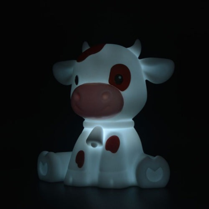 A cow-shaped night light glowing with a soft white light, featuring brown spots and a pinkish snout, set against a solid black background.