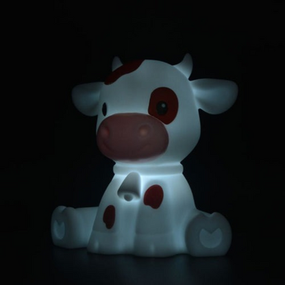 A cow-shaped night light glowing with a soft white light, featuring brown spots and a pinkish snout, set against a solid black background.