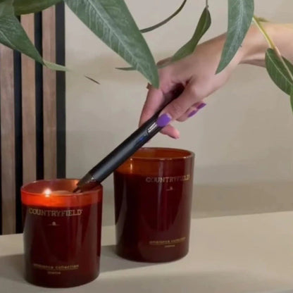 Two amber glass candles on a shelf, one lit and one unlit, with a hand holding a black lighter near the flame. 