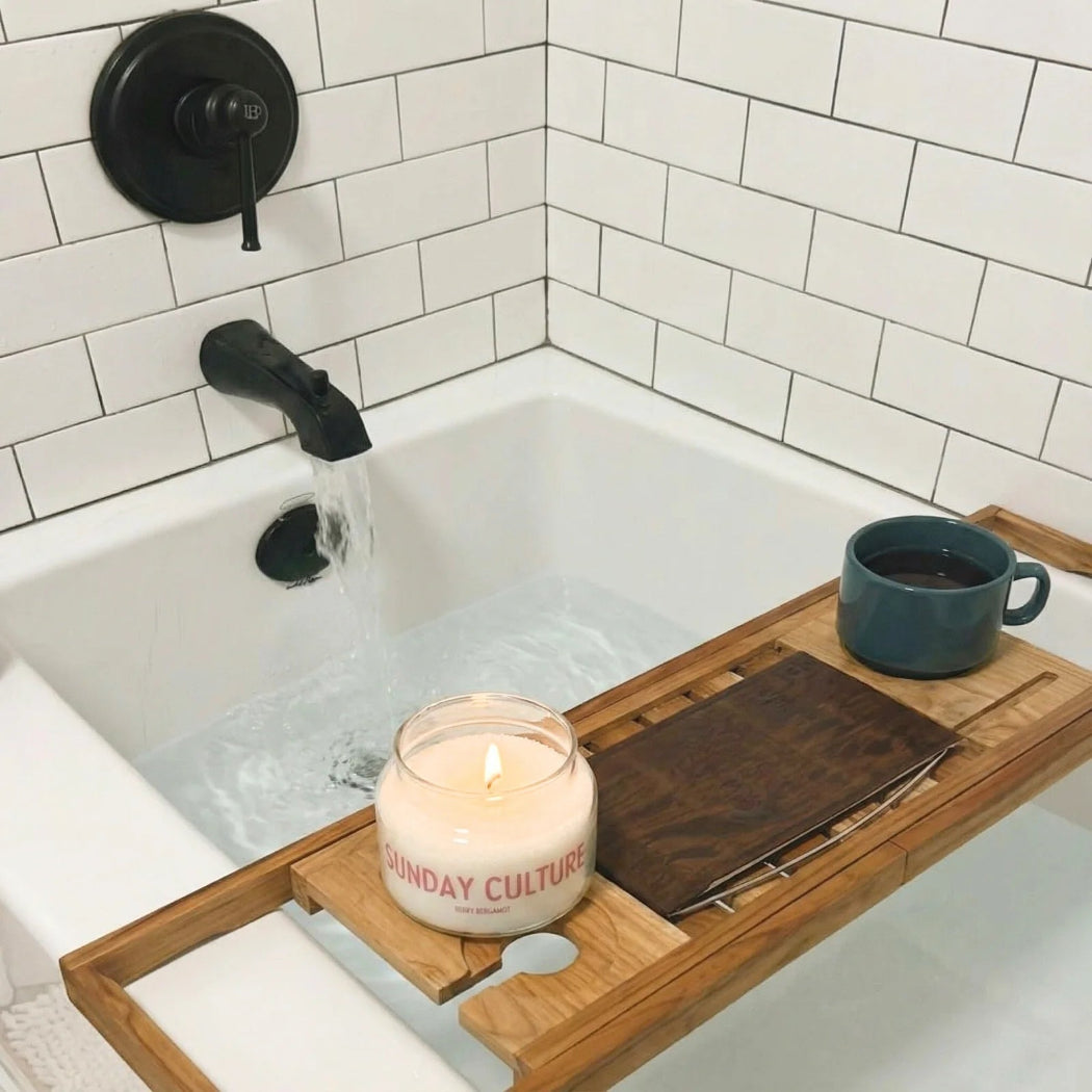 A wooden bath caddy draped over a white bathtub with running water, holding a lit candle labeled 'Sunday Culture', a dark brown book or journal, and a blue mug. The background shows a white subway-tile wall and a black faucet.