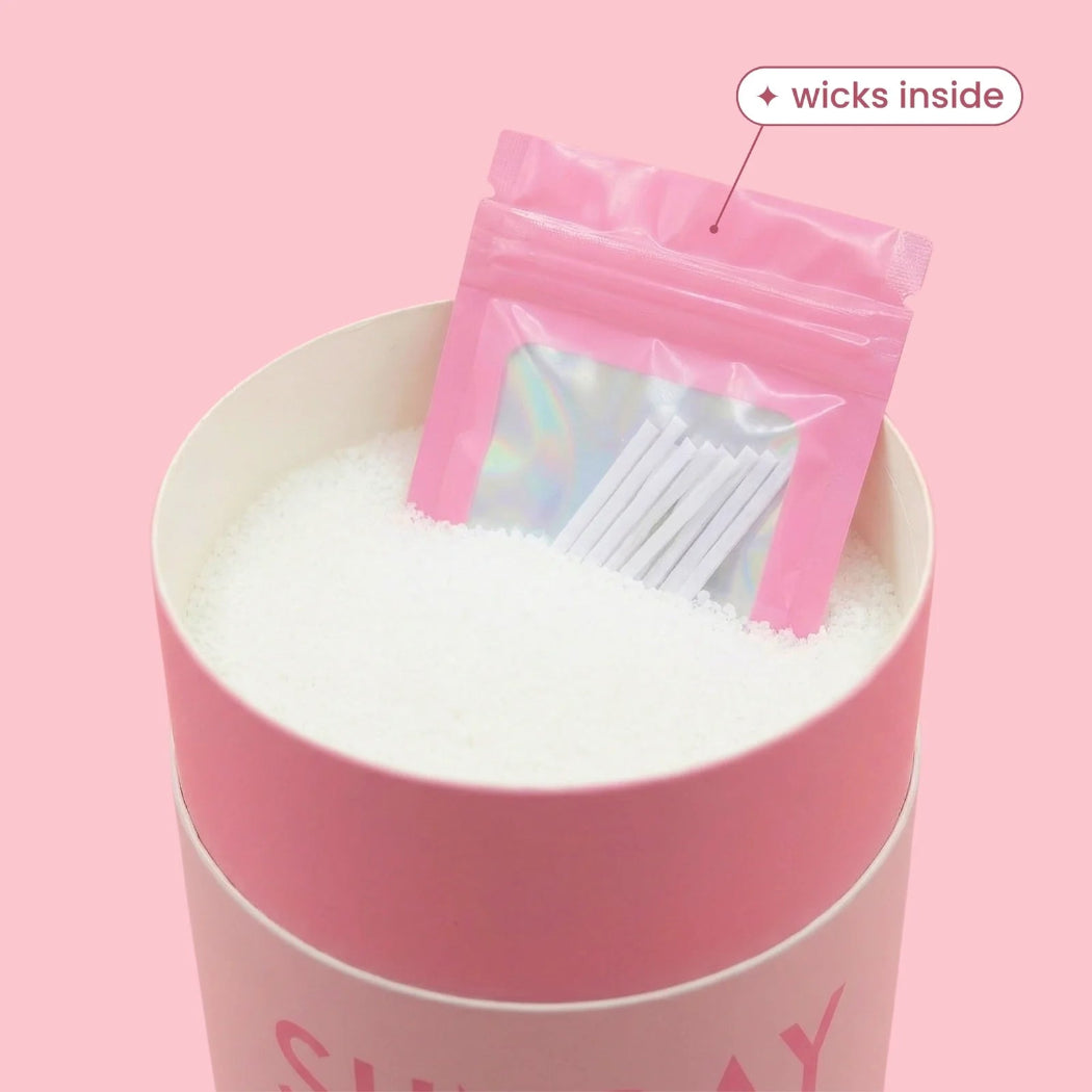 A pink cylindrical container filled with white granules, with a small pink resealable pouch labeled 'wicks inside' placed on top. The pouch has a holographic window showing white wicks.