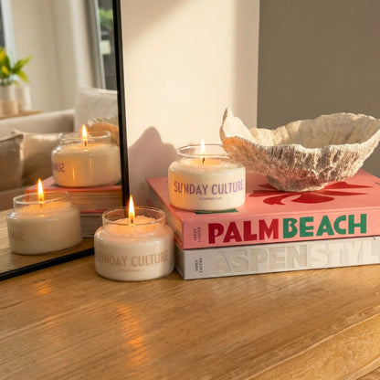 Three lit soy candles in clear glass jars with white wax and gold lettering reading 'SUNDAY CULTURE' are arranged on a wooden surface next to a mirror. The candles are styled on top of books, including one with 'PALM BEACH' on the cover, with a seashell bowl nearby.