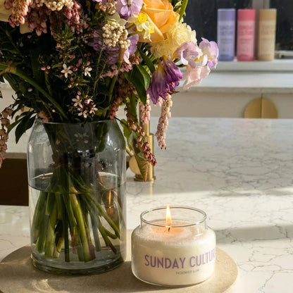 Candle with 'Sunday Culture' branding next to a vase of flowers on a marble surface.