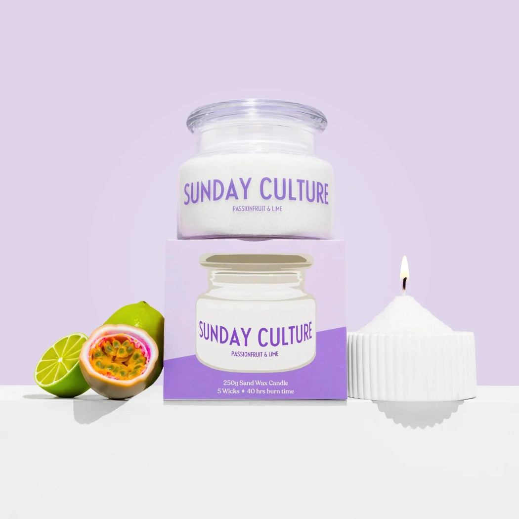 A white wax candle in a ribbed white vessel with a purple flame, shown next to its purple and white box and a glass jar candle labeled 'SUNDAY CULTURE PASSIONFRUIT & LIME' with passion fruit slices on a light lavender background.