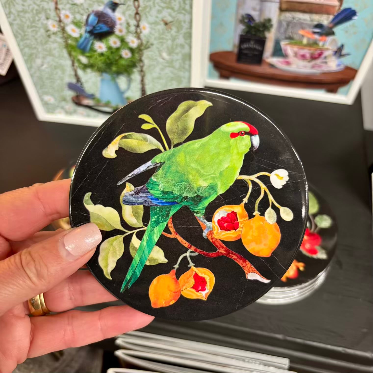 Hand holding a round coaster with a kakariki design against a blurred background