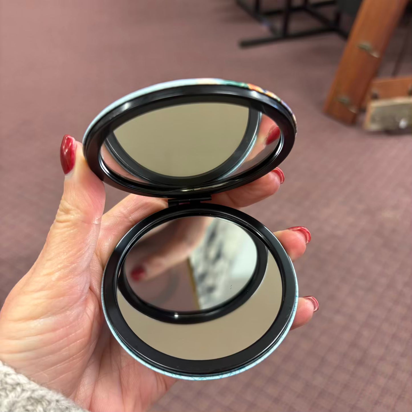 Compact mirror held by a hand with red nail polish on a blurred background