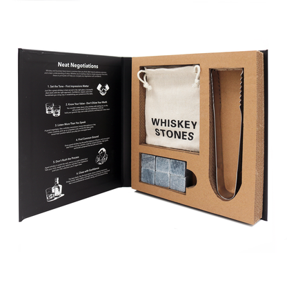 A whiskey stone set presented inside an open book-shaped box. The set includes a beige drawstring pouch printed with 'WHISKEY STONES', a metal tongs tool, and several gray stone cubes.