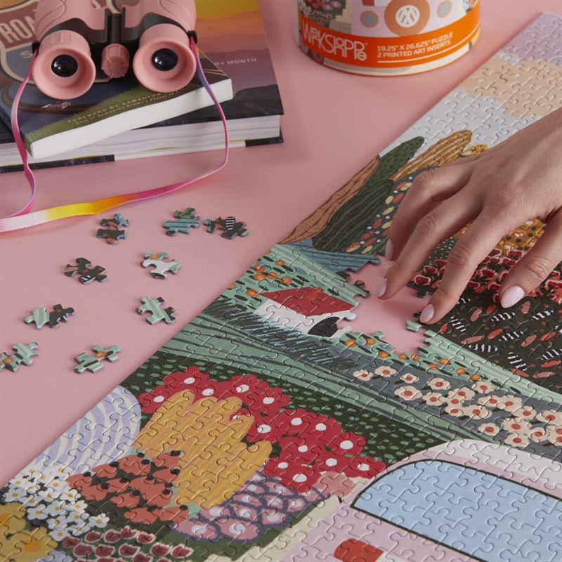 A person's hand placing a jigsaw puzzle piece on a colorful landscape puzzle, with the pink puzzle box and loose puzzle pieces nearby on a pink surface.