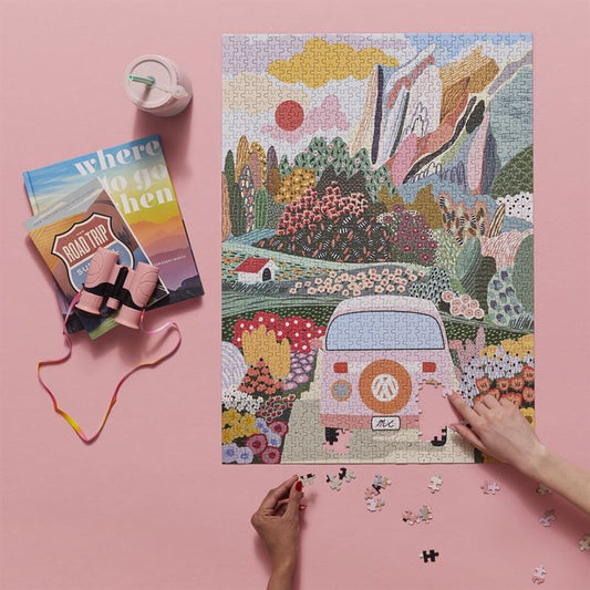 Jigsaw puzzle with a camper van design on a pink background, with books and a pencil nearby.