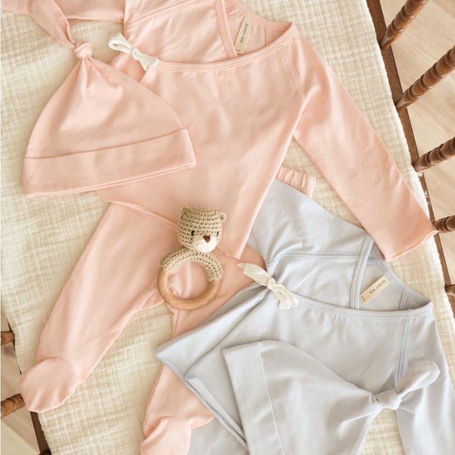 Baby clothing and accessories including a pink outfit, hat, and gray pants on a textured surface.