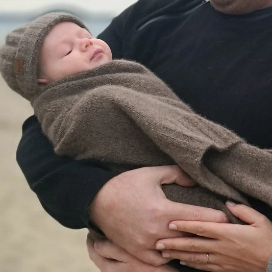 A sleeping newborn baby wrapped in a soft taupe knit blanket and wearing a matching knit beanie, held by an adult on a beach.