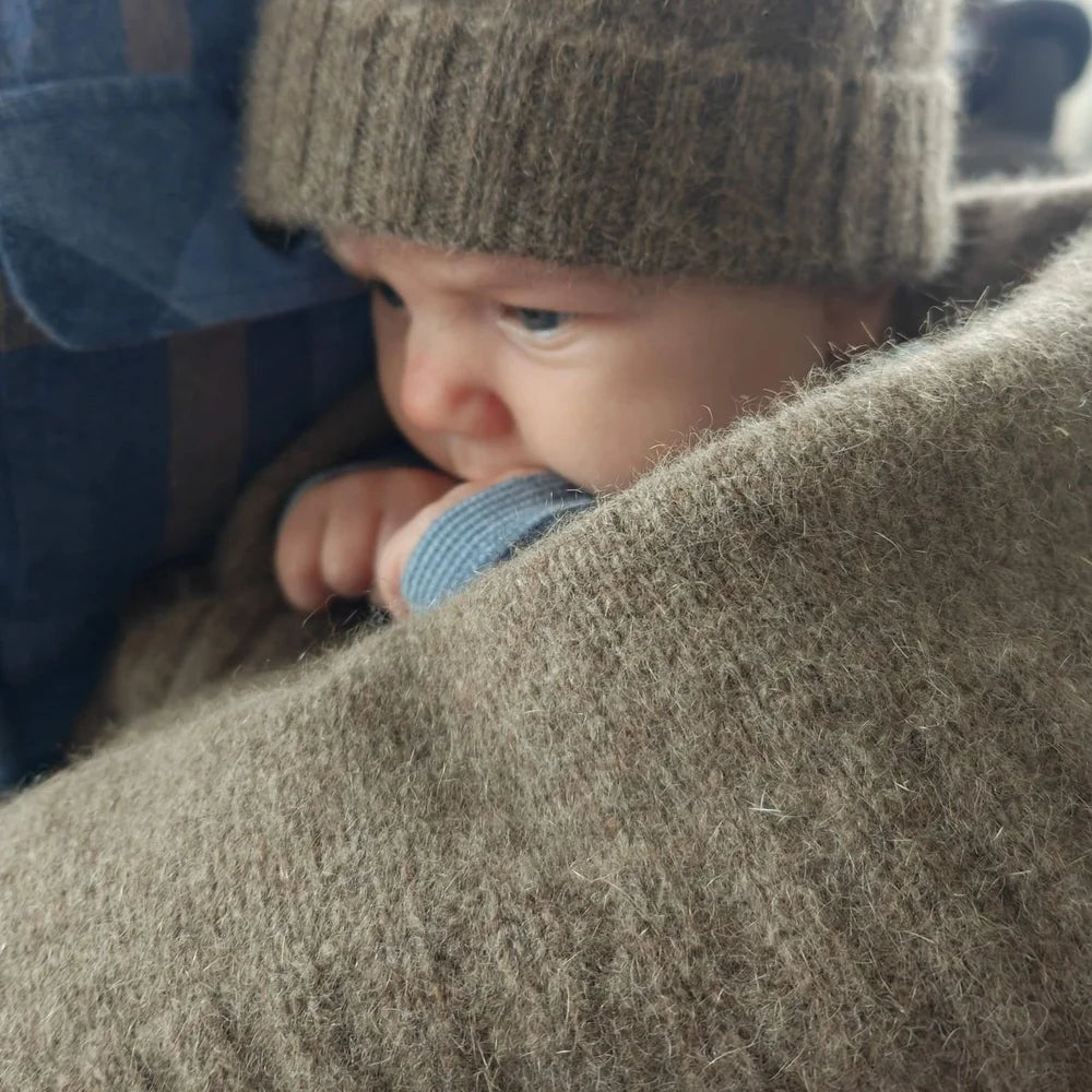 A close-up lifestyle shot of a baby wearing a brown knit beanie and a blue striped shirt, nestled in a cozy, soft brown wool blanket.