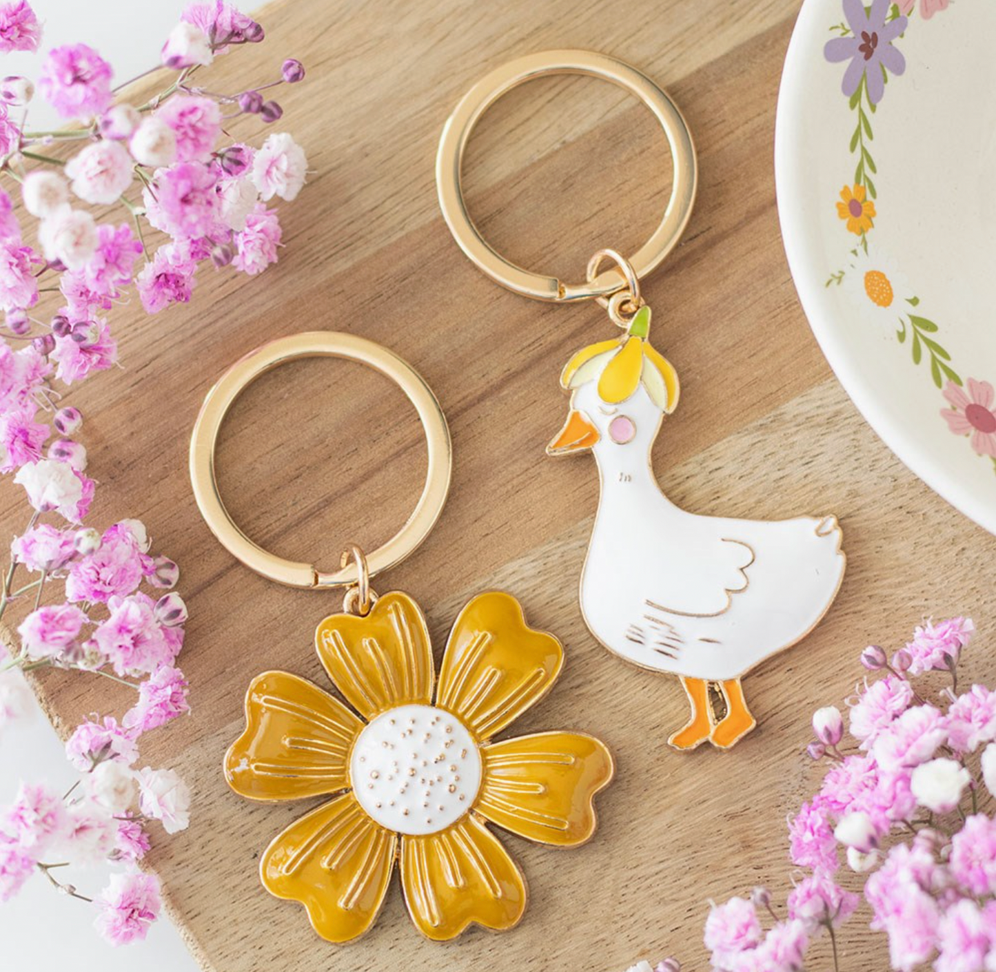 Two keychains, one with a floral design and the other with a goose, on a wooden surface with pink flowers.