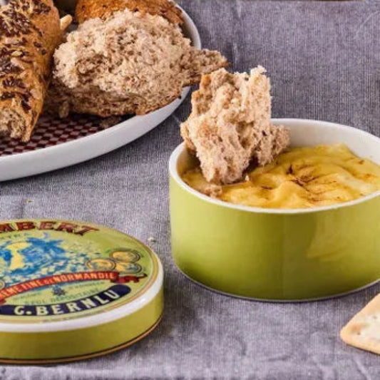 A round green ceramic bowl filled with yellow cheese and topped with a piece of rustic bread, set on a gray tablecloth. A matching green tin lid labeled C.