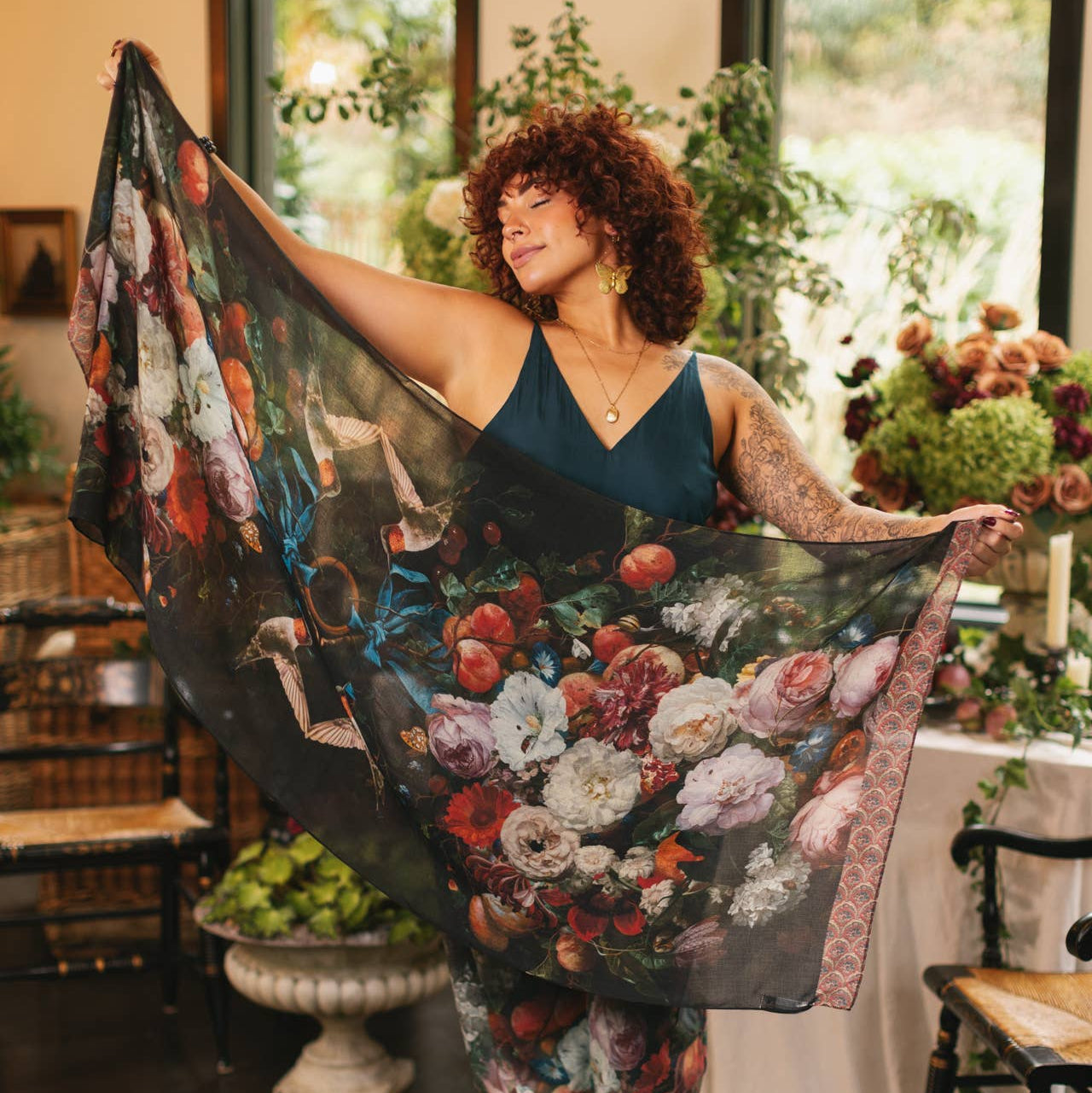 Woman holding a large floral-patterned scarf in a decorated room with plants and furniture.