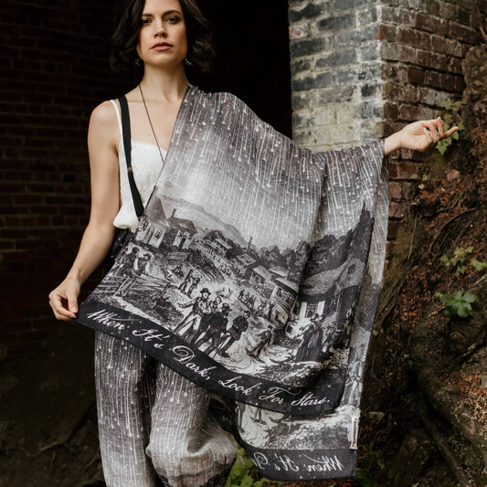Woman holding a large black and white patterned scarf against a brick wall.
