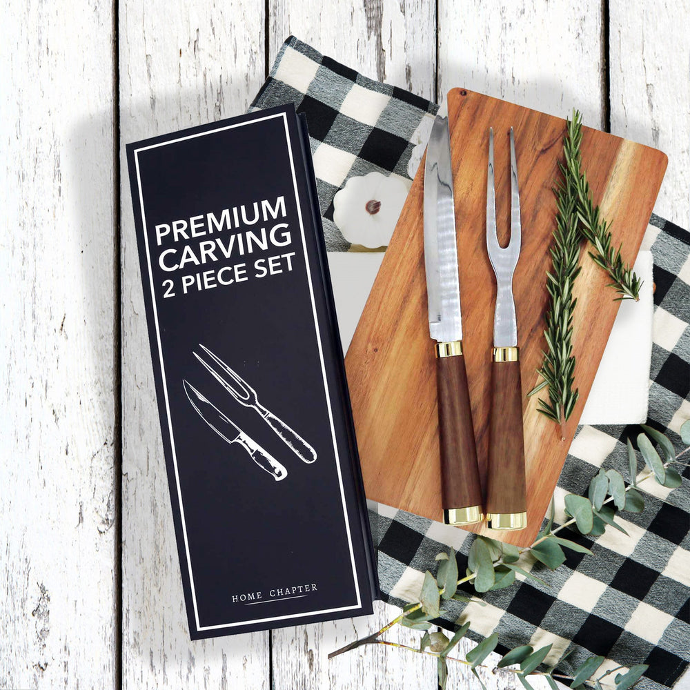Premium carving 2-piece set by Home Chapter, including a carving knife and carving fork, shown with their black presentation box on a rustic white wood background with a black-and-white checkered cloth, wooden cutting board, and greenery.