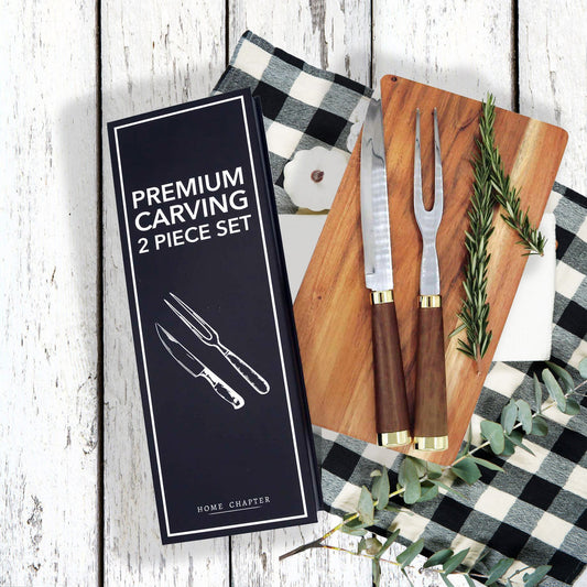 Premium carving 2-piece set by Home Chapter, including a carving knife and carving fork, shown with their black presentation box on a rustic white wood background with a black-and-white checkered cloth, wooden cutting board, and greenery.