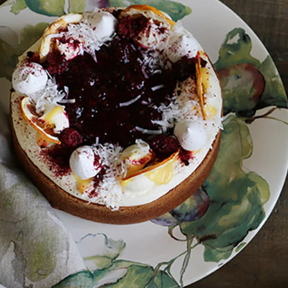 A round fruit and cream tart on a floral ceramic plate, set on a dark wooden table with a small white pitcher nearby.