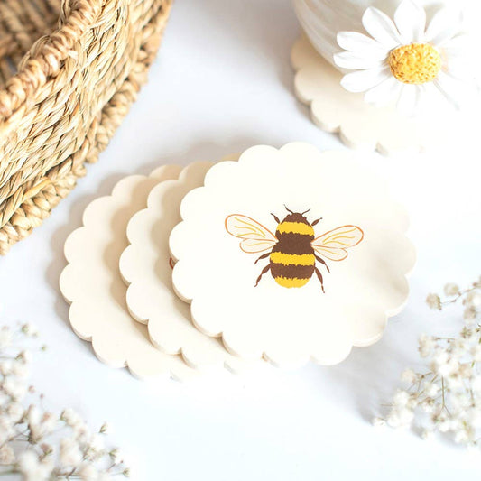 A stack of scalloped-edge cream ceramic coasters featuring a printed bumblebee design on the top coaster, styled with a woven basket, daisies, and baby's breath on a white surface.