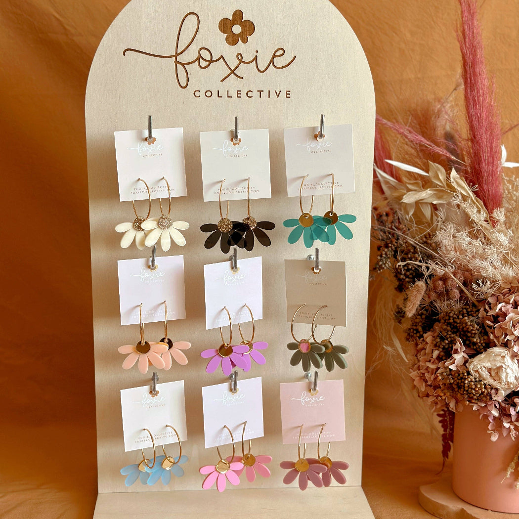 A display stand featuring multiple pairs of small flower-shaped earrings in assorted colors, branded with 'foxie collective' on the arch-shaped wooden board. The earrings are shown against a warm tan fabric backdrop with dried floral decor on the right.