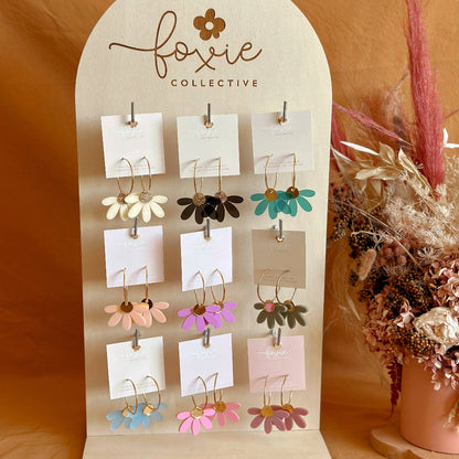 A display stand featuring multiple pairs of small flower-shaped earrings in assorted colors, branded with 'foxie collective' on the arch-shaped wooden board. The earrings are shown against a warm tan fabric backdrop with dried floral decor on the right.