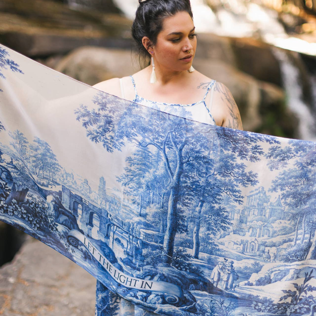 Woman holding a large blue and white scenic scarf in front of a waterfall.
