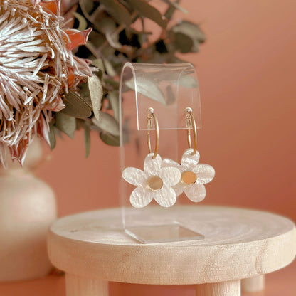 Flower-shaped earrings on a clear stand with a wooden tray and decorative plants on a peach-colored background