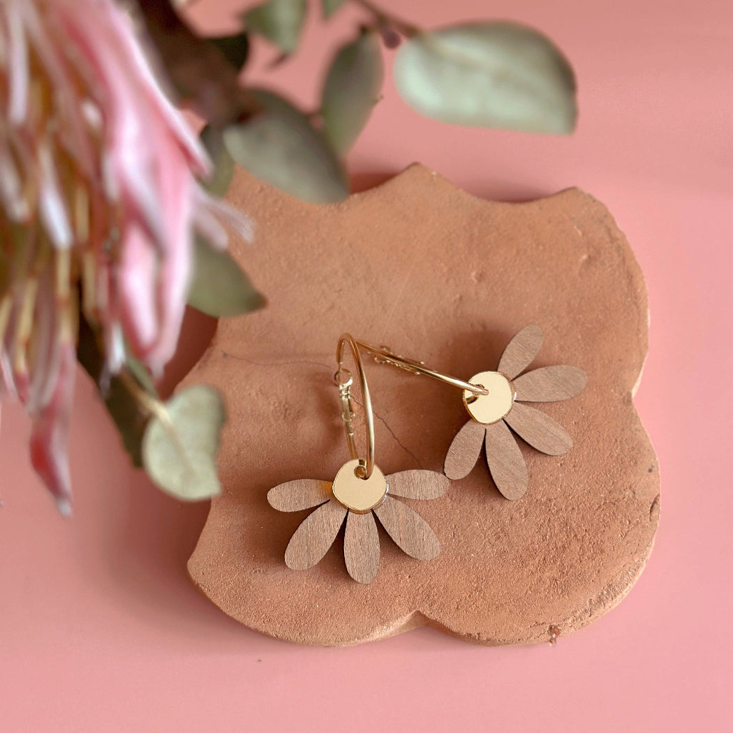 Gold flower-shaped earrings on a pink stone with pink flowers and green leaves in the background