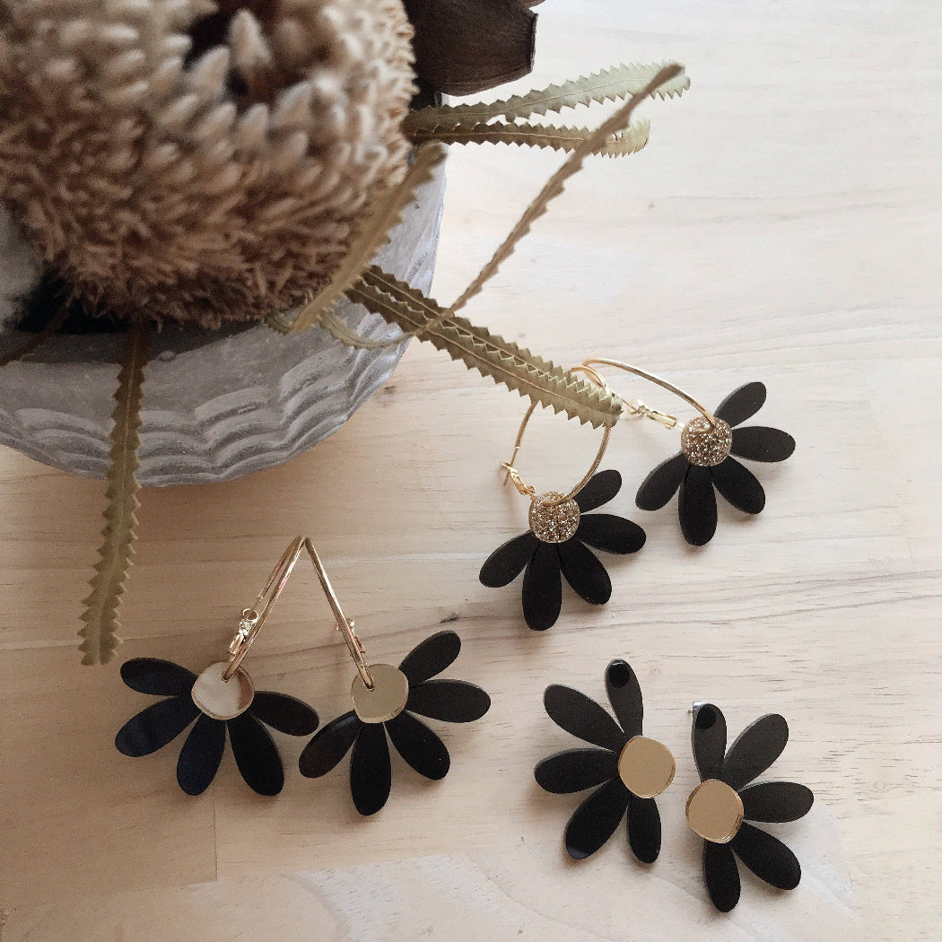 Black flower-shaped earrings on a light wooden surface with a decorative vase in the background.