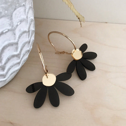 Black daisy flower hoop earrings with gold-tone metal hoops and discs, displayed on a light wood surface next to a textured white ceramic object.