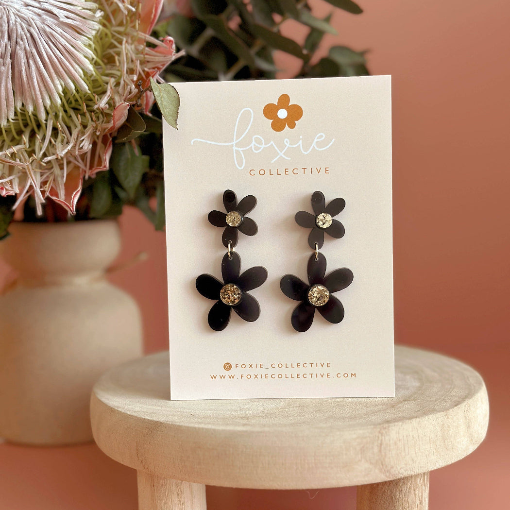 Black flower drop earrings with gold-tone center details, displayed on a white Foxie Collective card, set on a small wooden stand against a peach background with dried flowers.