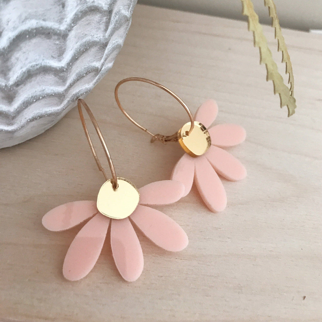 A pair of daisy flower hoop earrings with light pink petal charms and gold-tone hoops and centers, displayed on a light wood surface next to a textured gray ceramic object.
