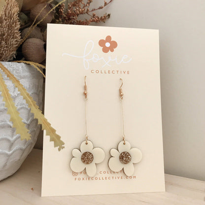 A pair of dangling flower earrings displayed on a cream card from Foxie Collective. The earrings have white flower-shaped charms with a glittery gold center and gold-tone hooks, set against a styled background with dried plants.