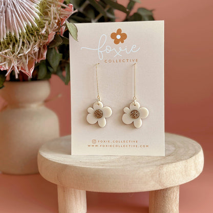 A pair of white flower dangle earrings with gold-tone centers and hooks, displayed on a white 'foxie collective' card, set on a small wooden stool against a dusty pink background with dried flowers.