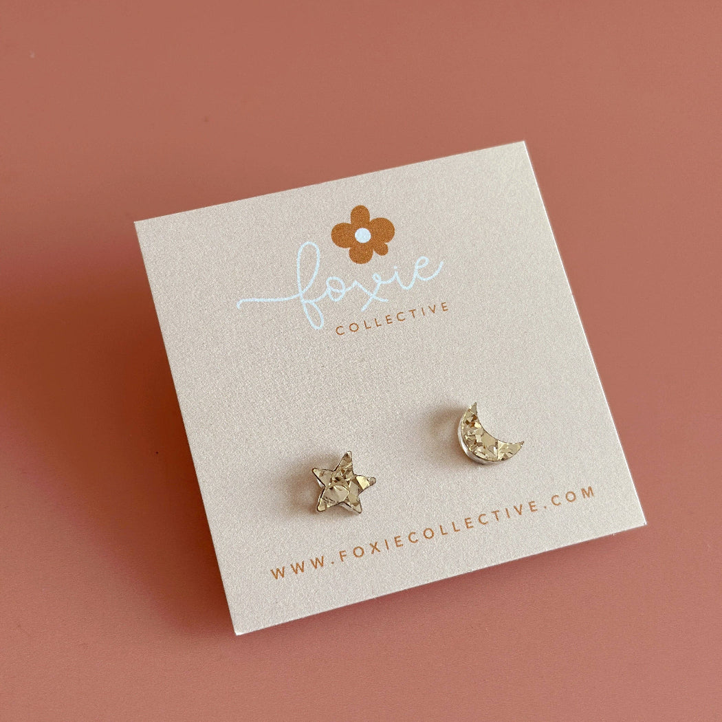 Pair of gold earrings on a 'Foxie Collective' branded card against a pink background