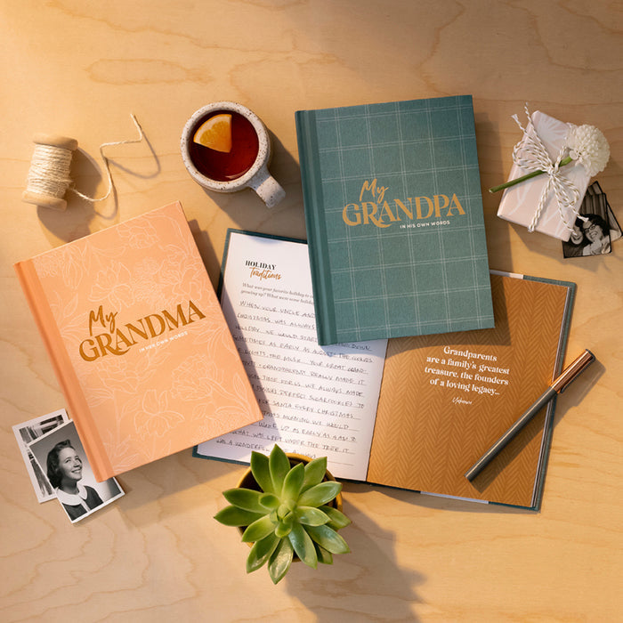 A styled flat-lay of two personalized hardcover books titled 'My Grandma' and 'My GRANDPA' on a light wood surface, surrounded by tea, a succulent, a small photo, thread, and wrapped gift with string.